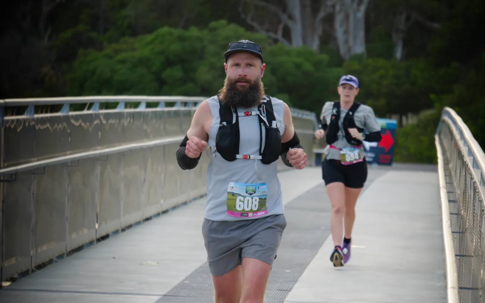 Two runners crossing a steel footbridge at the Coastal Pathway Ultra, a bearded runner in front