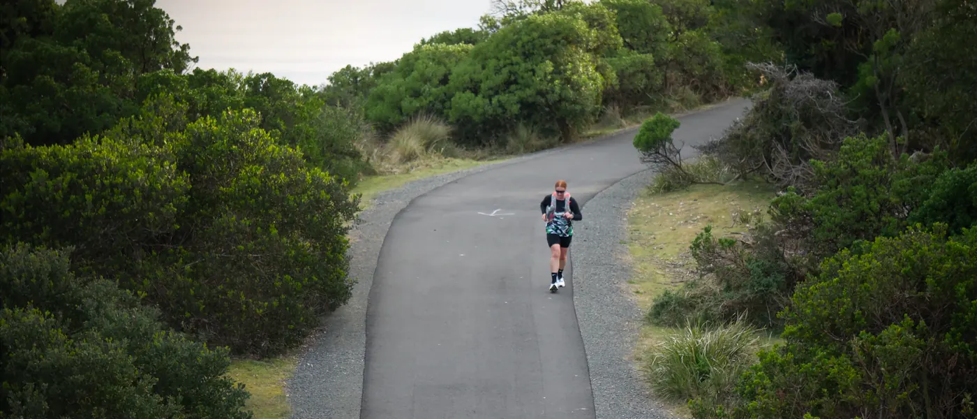 A runner alone on a sealed coastal path curving through dense scrub, with water and a headland behind