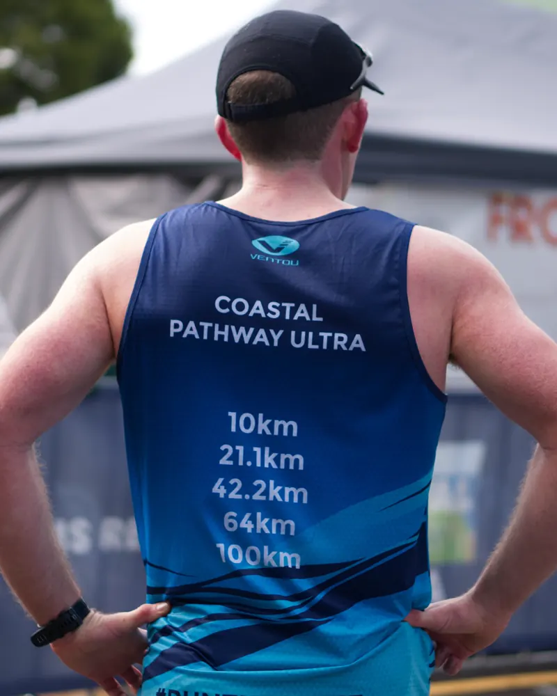 A runner seen from behind, hands on hips, in a Coastal Pathway Ultra singlet listing the race distances