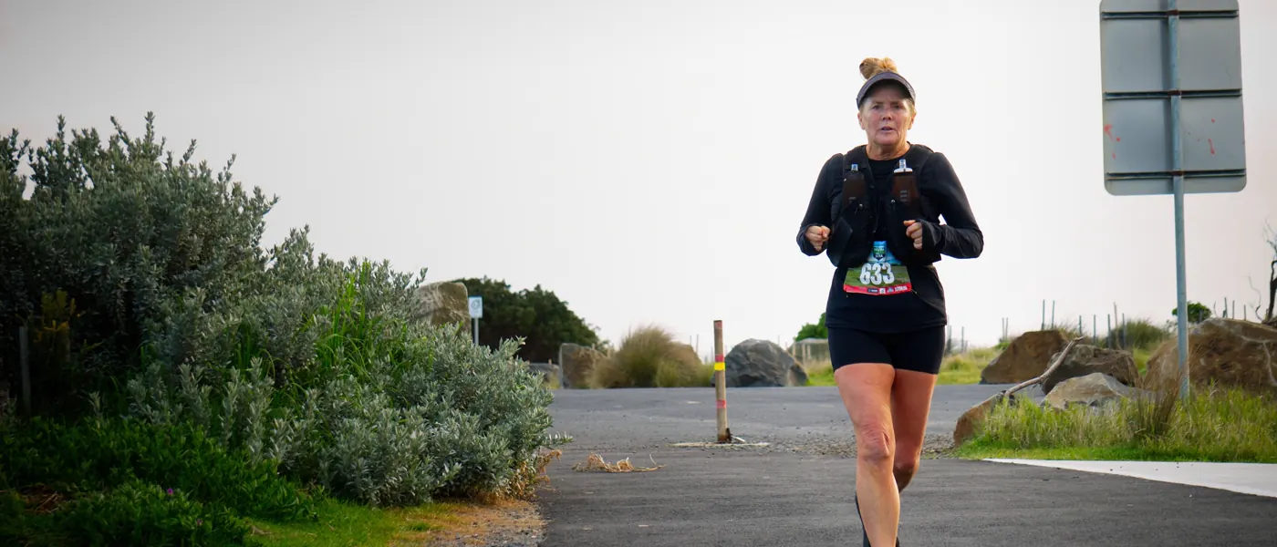 A runner in a race vest on a sealed coastal path under a pale sky, scrub on the left, rocks and grass on the right